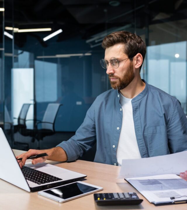 A focused professional working at a desk with a laptop, documents, and a calculator in a modern office setting.