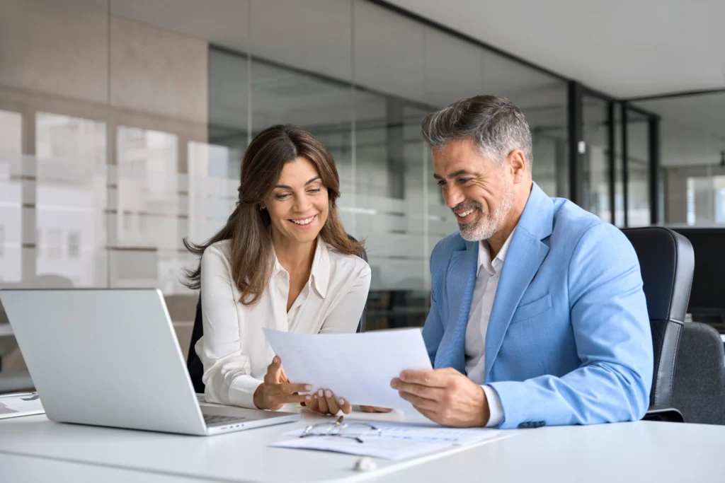 Two happy busy middle aged professionals man and woman business leaders partners checking document reading financial report talking working together on laptop computer in office at corporate meeting.